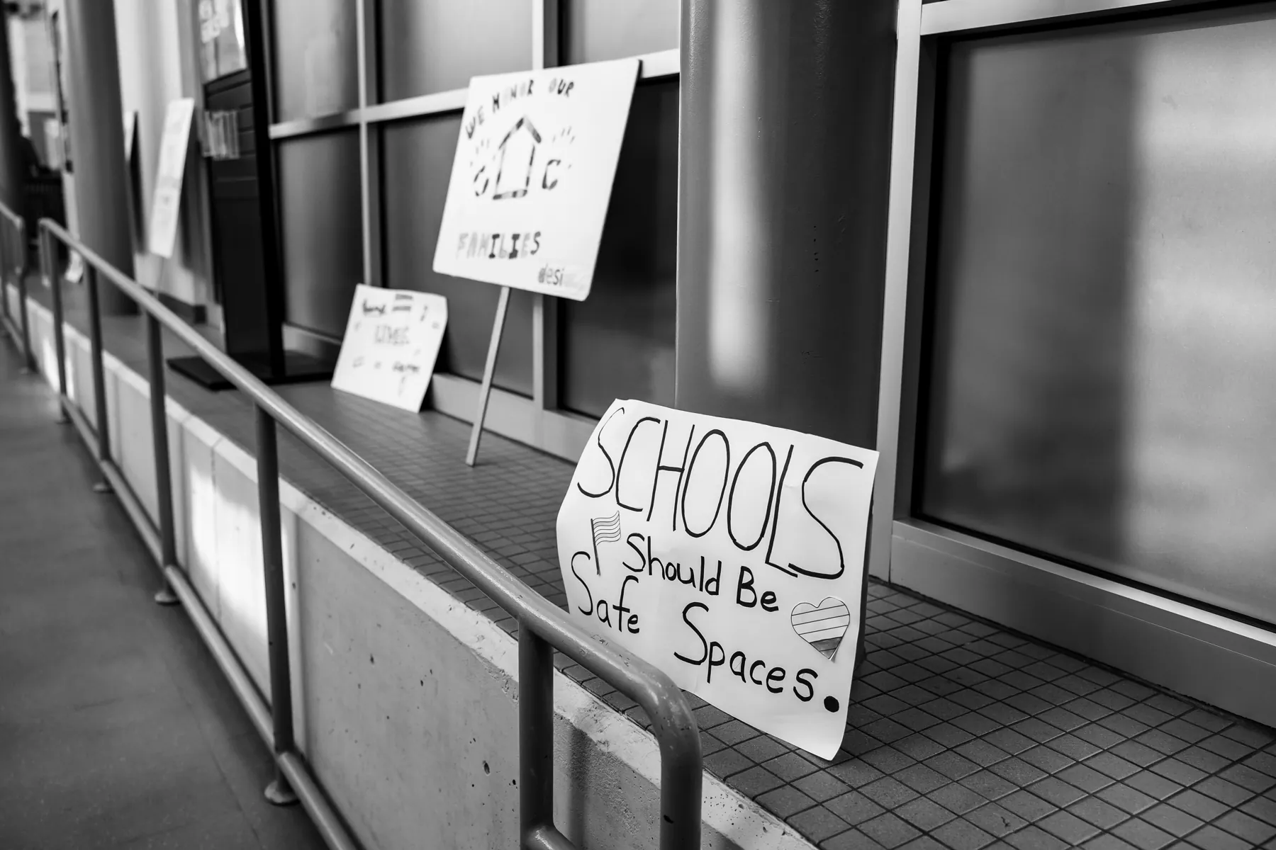 Black-and-white photo of handmade signs propped along a window ledge at the Desi Rainbow event in Edison, New Jersey, one reading Schools should be safe spaces and another We honor our families.