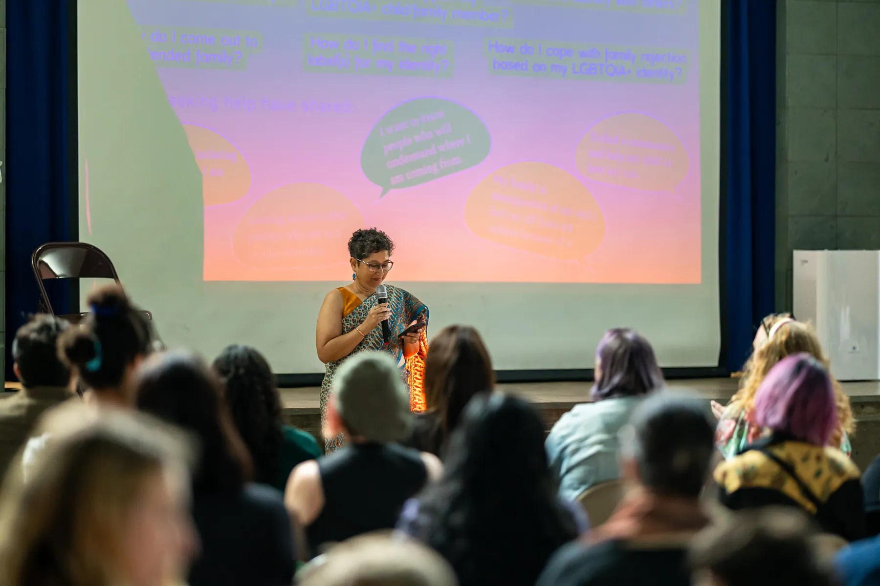A speaker in a sari with a microphone presents to a seated audience at the Desi Rainbow event with Alok Vaid-Menon in Edison, New Jersey, in front of a projected slide of questions LGBTQIA+ people ask.