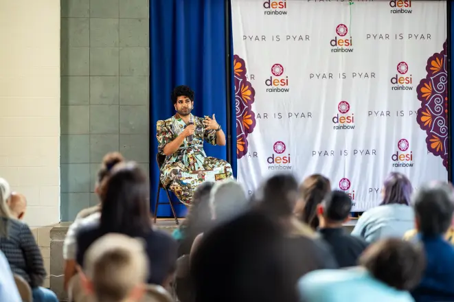 Alok Vaid-Menon speaking on stage in front of a Pyar is Pyar backdrop to a seated audience in Edison, New Jersey.