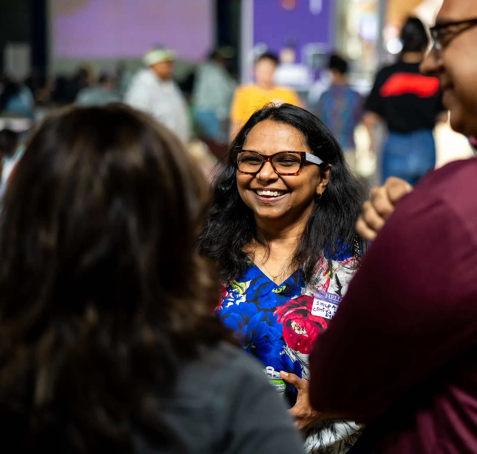 A smiling woman in glasses and a floral top, wearing a Hello name tag, talks with two people at a busy Desi Rainbow event in Edison.