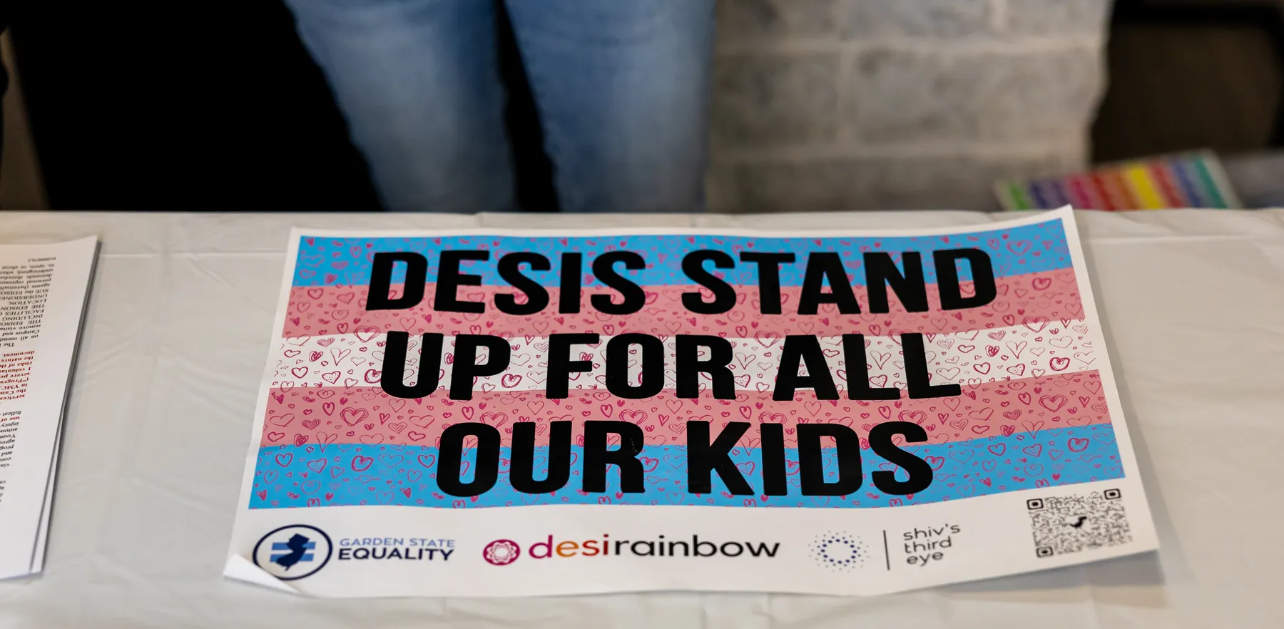 The welcome table at the Desi Rainbow event with Alok Vaid-Menon in Edison, New Jersey: a trans-flag sign reading Desis stand up for all our kids, with the Garden State Equality, Desi Rainbow and Shiv’s Third Eye logos.