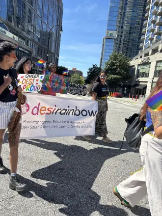 Marchers carrying a Desi Rainbow banner and a Queer Desi Proud sign down a city street at Atlanta Pride.