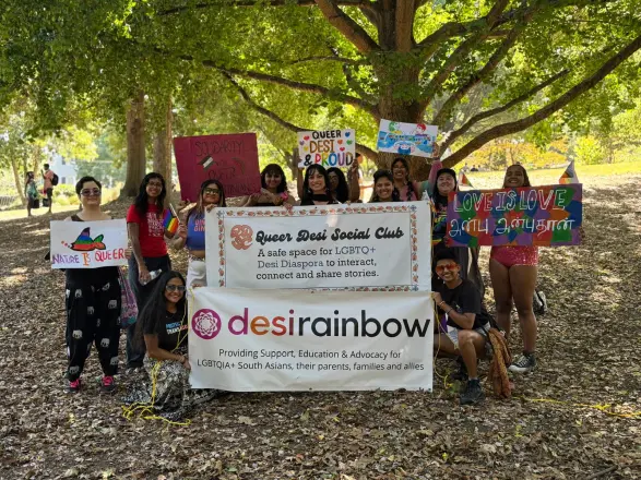 A group under a tree at Atlanta Pride with Queer Desi Social Club and Desi Rainbow banners and hand-made signs.