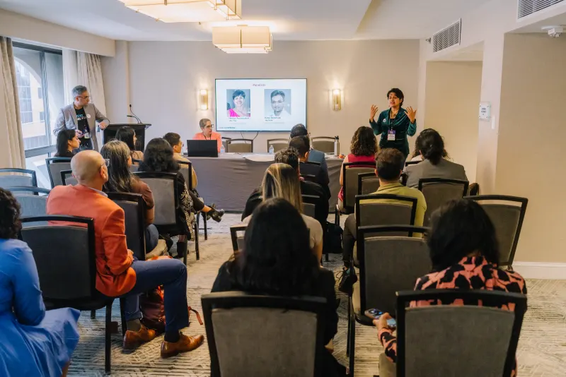 A speaker addresses a seated audience at the Breaking the Silence panel in Chicago, with photos of the panelists on a screen at the front of the room.
