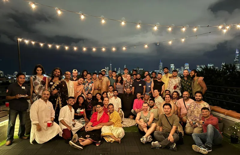 A large crowd of about fifty people, many in festive Indian clothing, gathered on a Chicago rooftop at night under string lights for an Onam potluck with Trikone Chicago, the city skyline behind them.