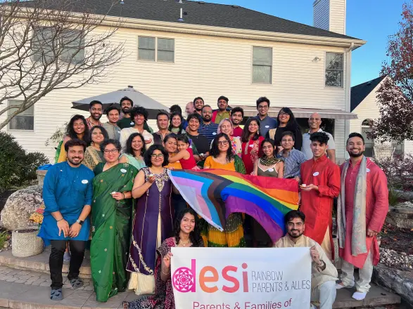 A large group in festive clothes celebrating Diwali 2022 outside a house, holding a rainbow flag and a Desi Rainbow banner.