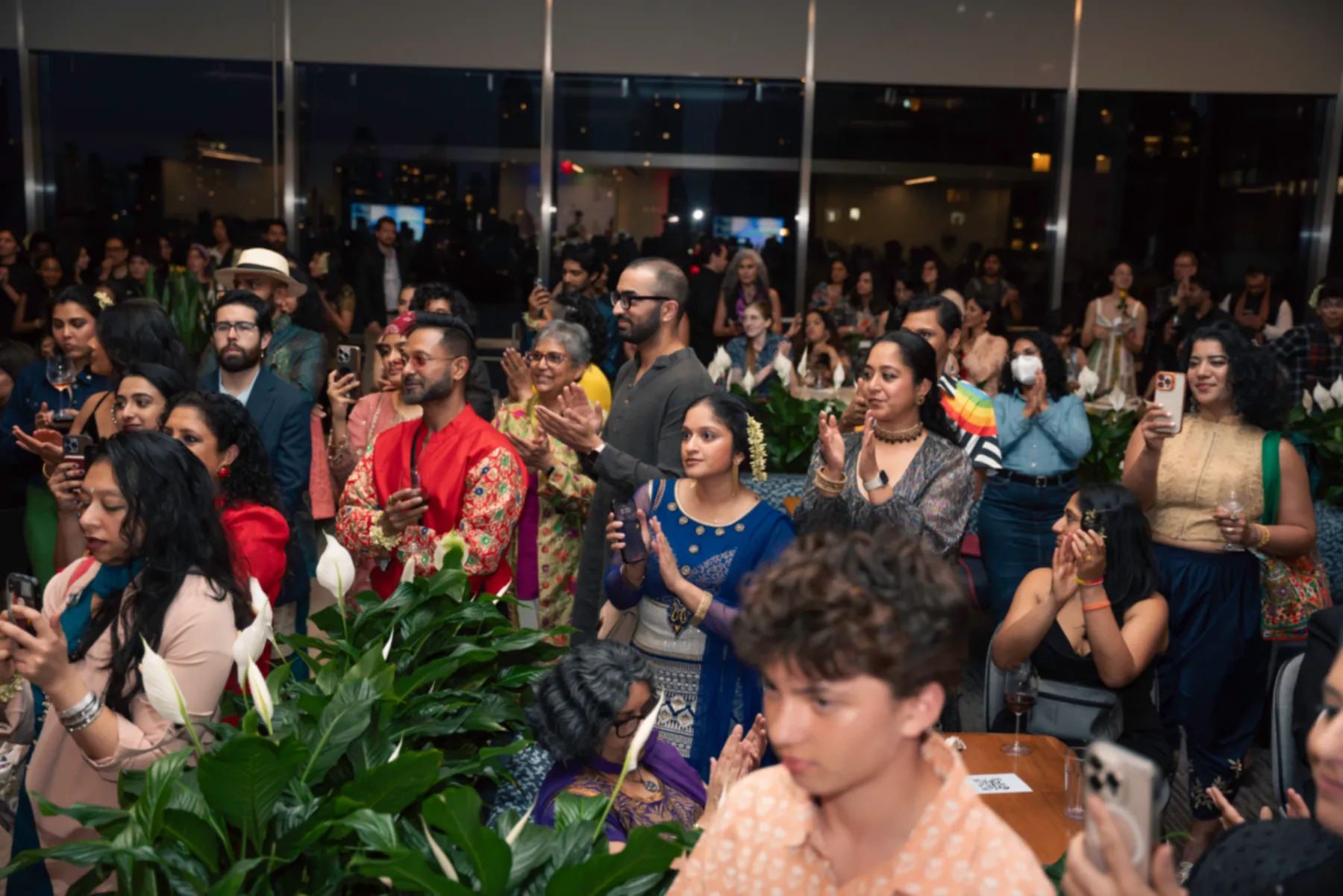 Guests standing and clapping at Desi Rainbow’s 2025 fundraiser, some filming on their phones.