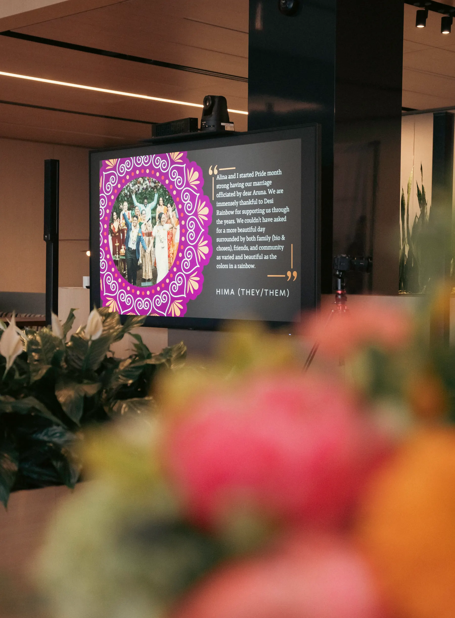 At Desi Rainbow’s 2025 fundraiser, a screen shows Hima’s story about their Pride-month wedding, officiated by Aruna, beside a photo of the wedding, with flowers blurred in the foreground.