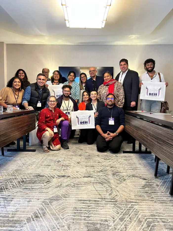 About twenty people posing together at a conference, several holding signs that read Proud to be a Desi Rainbow Ally.