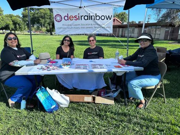 Volunteers sitting at a table under a Desi Rainbow tent on a sunny lawn.