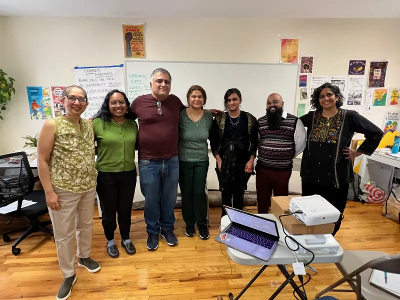 Seven people standing together in a community room after a Jahajee training, with community agreements and posters on the wall behind them.