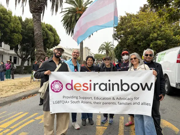 A group standing in the street behind a Desi Rainbow banner at Pride 2026, with a transgender pride flag above them.