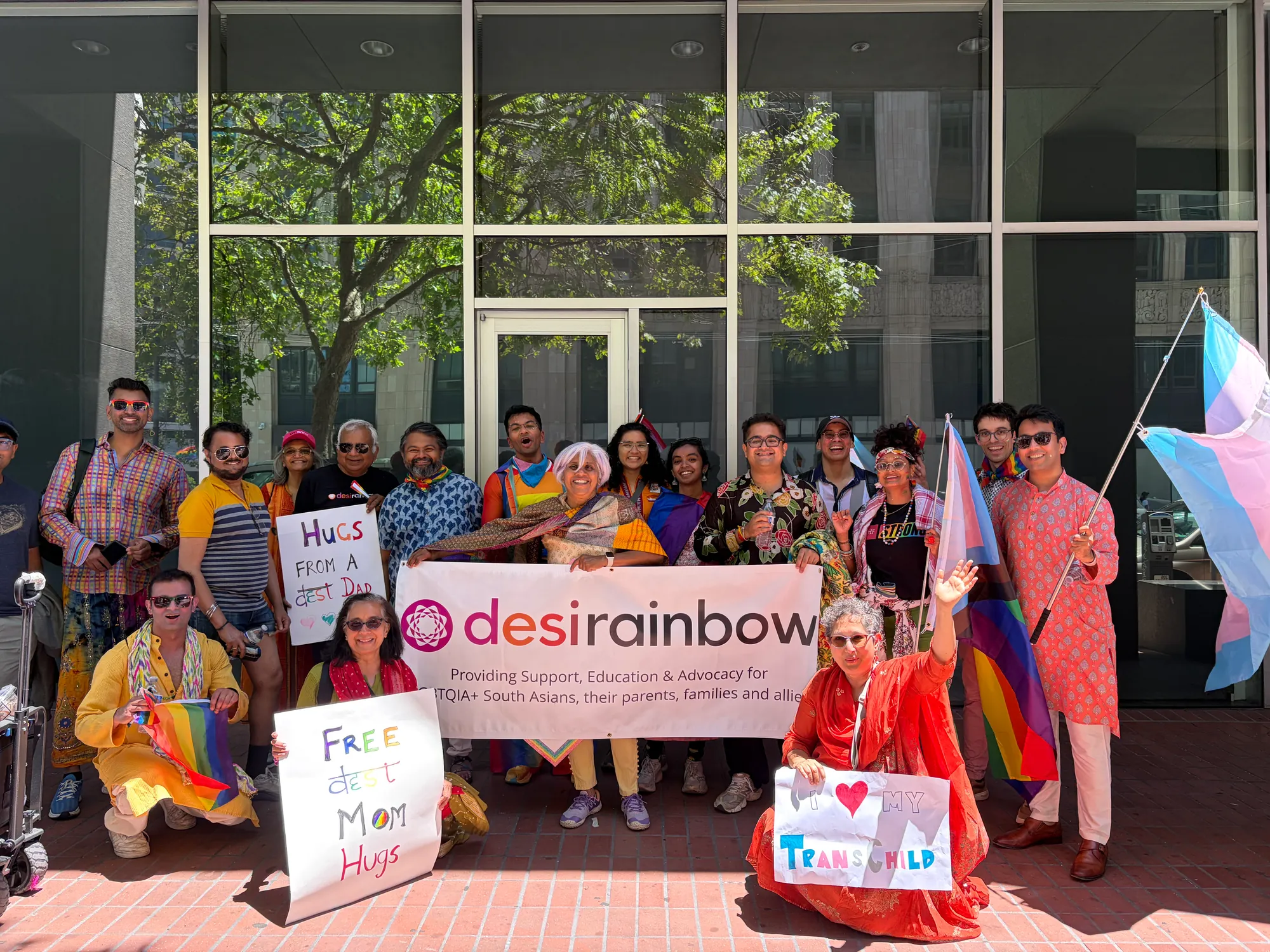 A big Desi Rainbow group at Pride in June 2026, gathered behind the Desi Rainbow banner with Pride and trans flags and handmade signs reading Hugs from a Desi Dad, Free Desi Mom Hugs, and I love my trans child.