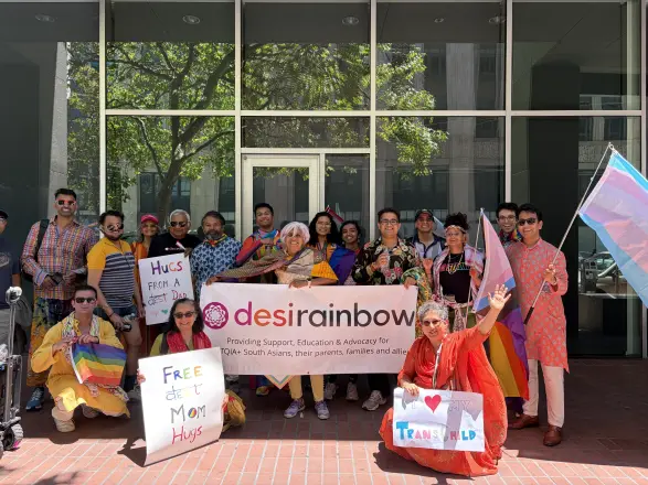 A big group gathered behind a Desi Rainbow banner with signs including Free Mom Hugs at Pride 2026.