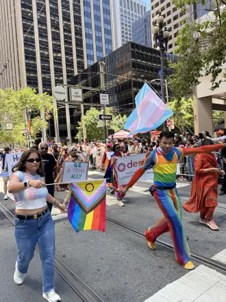Marchers in the Pride 2026 parade, one waving a Progress Pride flag and another in a rainbow outfit.
