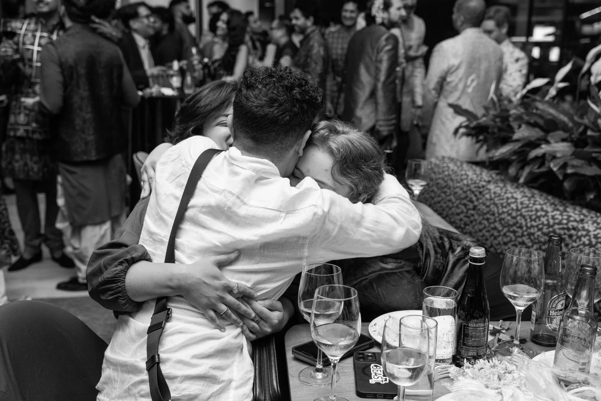 Black-and-white photo of friends in a long, close hug at a table during Desi Rainbow’s 2025 fundraiser, with guests talking in the background.