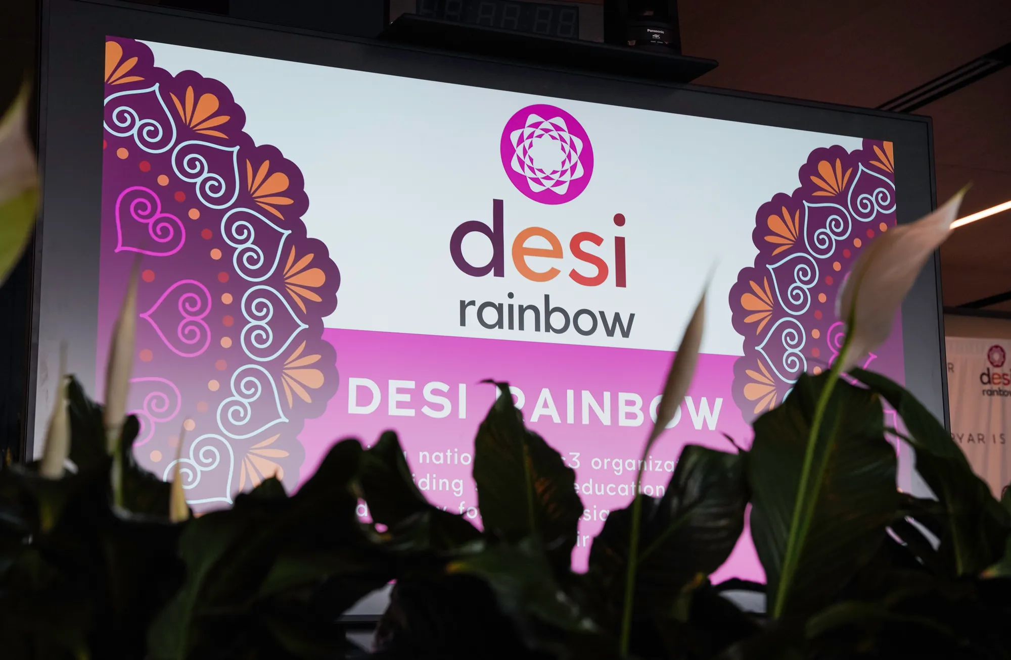The Desi Rainbow logo and mandala artwork on a big screen at Pyaar is Pyaar, seen through the leaves of plants in the foreground.
