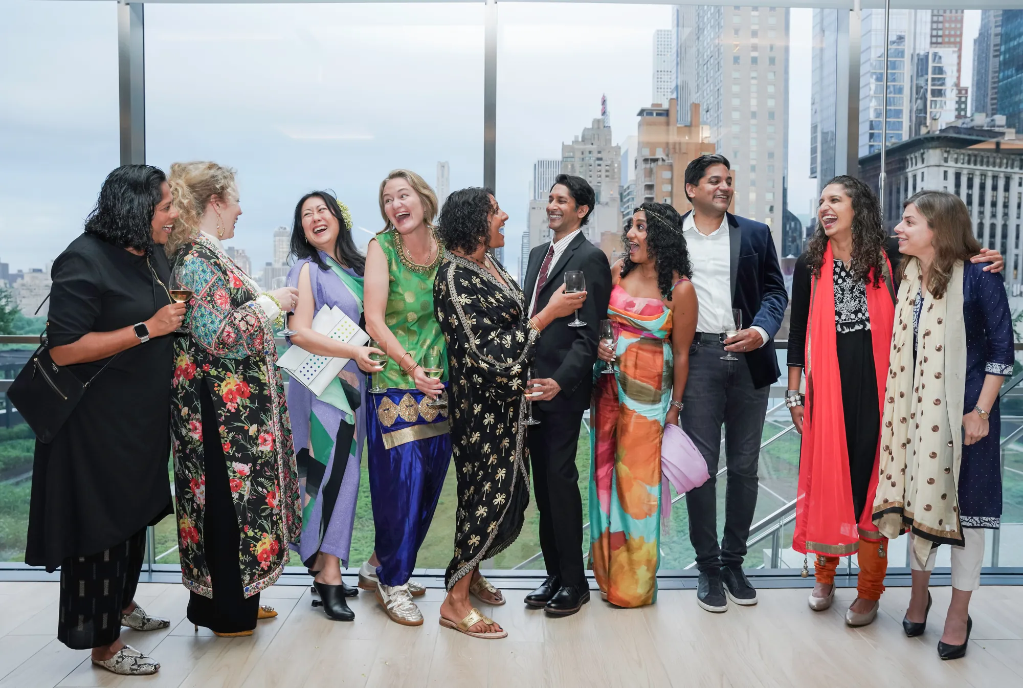 Eleven people in festive South Asian and formal outfits laughing together at Pyar is Pyar, in front of a wall of windows overlooking the New York City skyline.