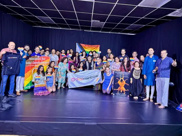 A large group posing together on a stage with rainbow flags and a Desi Rainbow banner.