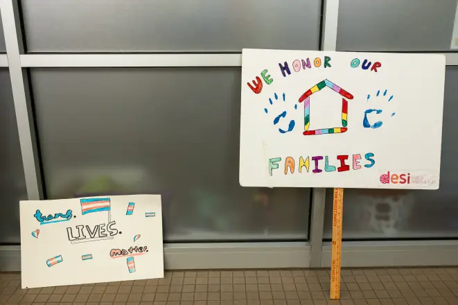 A hand-painted sign reading We Honor Our Families, with a rainbow house and a transgender pride flag.
