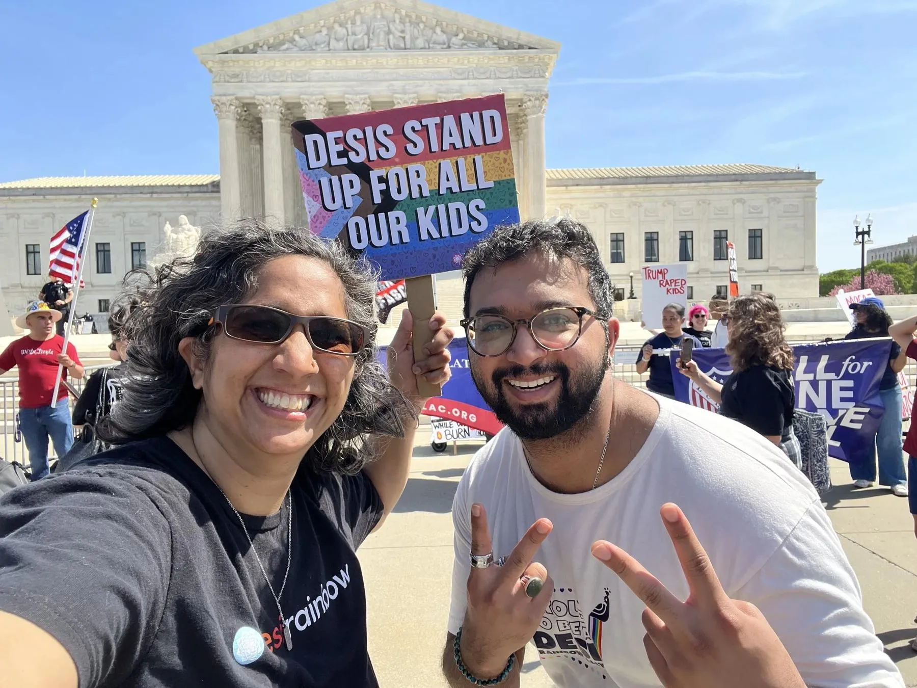 Two Desi Rainbow members smiling at an April rally outside the U.S. Supreme Court, one holding a rainbow sign reading Desis Stand Up For All Our Kids.