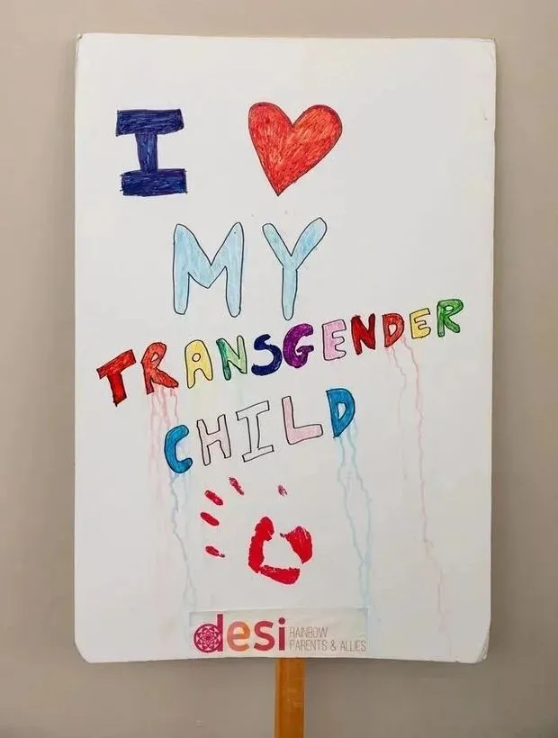 A hand-lettered sign reading I love my transgender child, with the Desi Rainbow logo at the bottom.