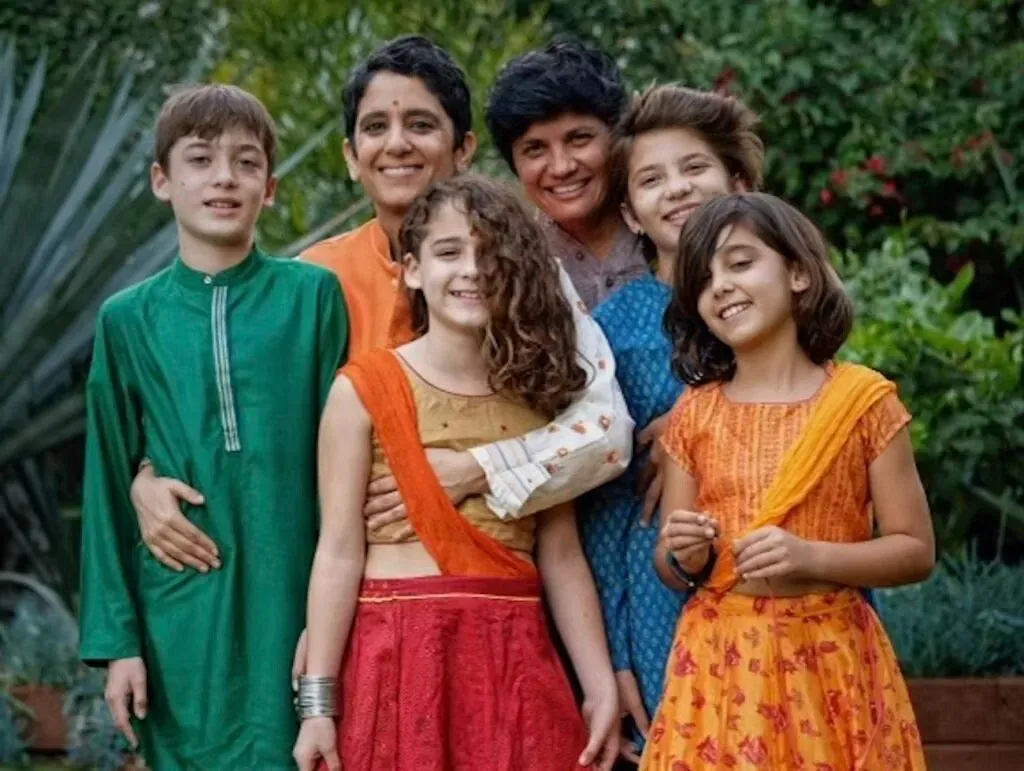 Two mothers and their four children, dressed in bright South Asian clothes, smiling together in a garden.